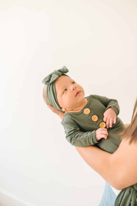 Baby Wearing Luna Luz Knotted Gown and Bow Headband in Olive Held by Mother